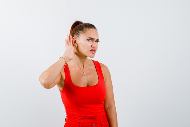 Mujer joven en camiseta roja, pantalones sosteniendo la mano detrás de la oreja y mirando enfocado, vista frontal.