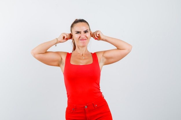 Foto gratuita mujer joven en camiseta roja, pantalones tirando de sus orejas y mirando divertido, vista frontal.