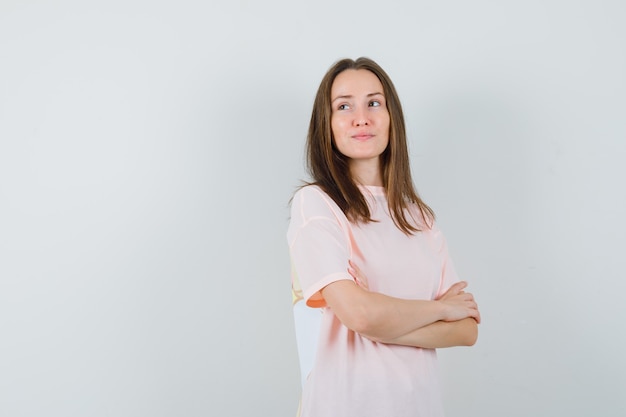 Mujer joven en camiseta rosa de pie con los brazos cruzados y mirando soñadora, vista frontal.