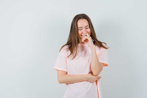 Mujer joven en camiseta rosa de pie en pose de pensamiento y mirando alegre, vista frontal.