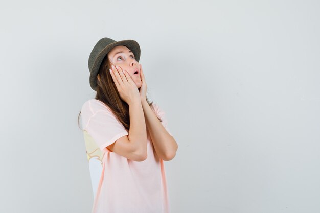 Mujer joven en camiseta rosa, sombrero cogidos de la mano en las mejillas y mirando asombrado, vista frontal.