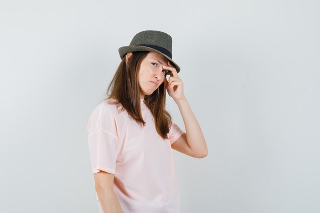 Mujer joven en camiseta rosa, sombrero frotándose las sienes y mirando triste, vista frontal.