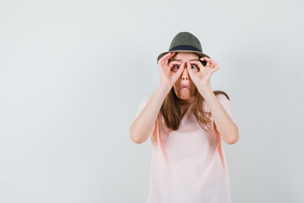 Mujer joven en camiseta rosa, sombrero mostrando gesto de gafas y mirando divertido, vista frontal.