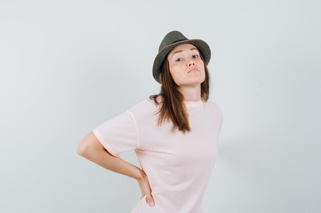 Mujer joven en camiseta rosa, sombrero que sufre de dolor de espalda y parece cansado, vista frontal.