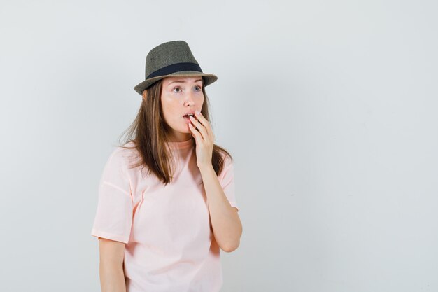 Mujer joven en camiseta rosa, sombrero sosteniendo la mano en la boca abierta y mirando sorprendido, vista frontal.