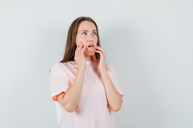 Mujer joven en camiseta rosa sosteniendo los dedos en las mejillas y mirando pensativo, vista frontal.