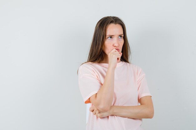 Mujer joven en camiseta rosa sosteniendo la mano en la barbilla y mirando pensativo, vista frontal.