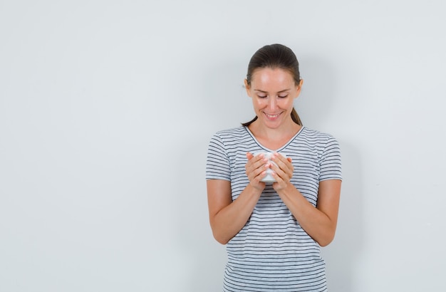 Mujer joven en camiseta sosteniendo una taza de té y mirando alegre, vista frontal.