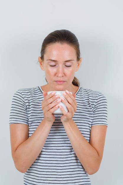 Mujer joven en camiseta sosteniendo una taza de té con los ojos cerrados, vista frontal.