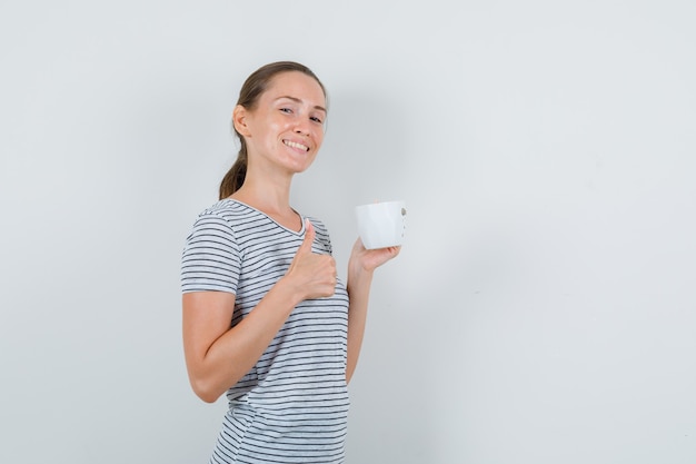 Mujer joven en camiseta sosteniendo una taza de té con el pulgar hacia arriba y mirando alegre, vista frontal.