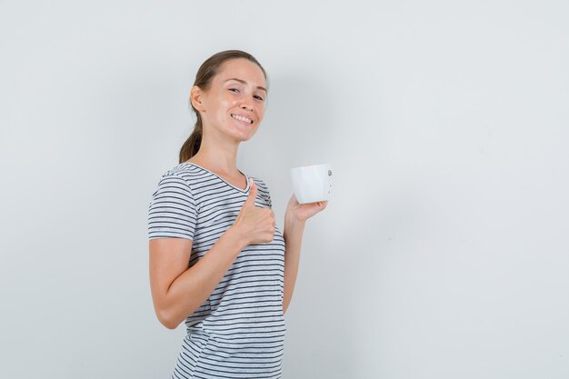 Mujer joven en camiseta sosteniendo una taza de té con el pulgar hacia arriba y mirando alegre, vista frontal.