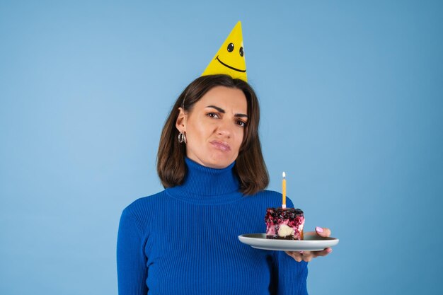 Mujer joven en un campo de golf en una pared azul celebra un cumpleaños, sostiene un trozo de tarta, es infeliz, decepcionada, no quiere envejecer