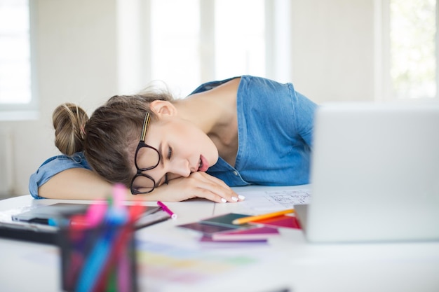 Mujer joven cansada con anteojos en la cabeza durmiendo en el escritorio mientras trabaja en la oficina moderna