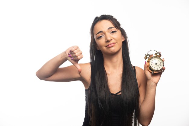 Mujer joven cansada con el pulgar hacia abajo sosteniendo un reloj de alarma en la pared blanca