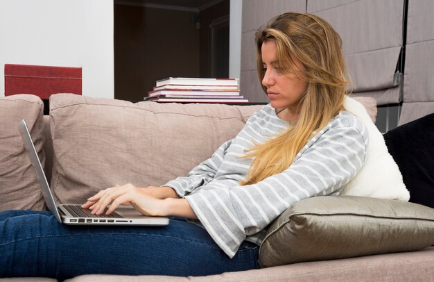 Mujer joven cansada que se sienta en el sofá usando el ordenador portátil