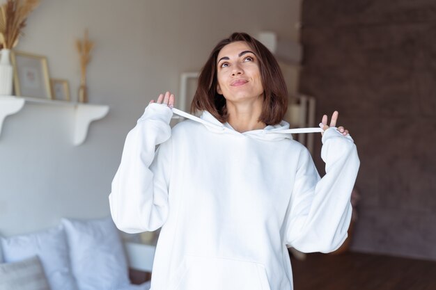 Mujer joven en casa en el dormitorio con una sudadera con capucha blanca cálida, feliz, posando, sonriente, acogedora noche de invierno