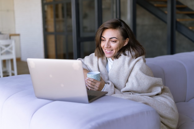 Mujer joven en casa en un sofá en invierno bajo una acogedora manta con un ordenador portátil, viendo series de comedia