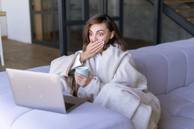 Mujer joven en casa en el sofá en invierno bajo una manta acogedora con una computadora portátil, mira sorprendida a la pantalla