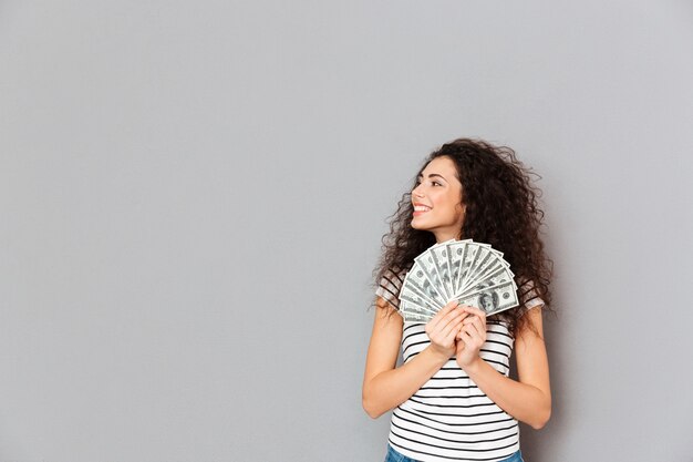 Mujer joven en casual holding fan de billetes de 100 dólares en las manos mirando a un lado con una amplia sonrisa alegrarse por la pared gris