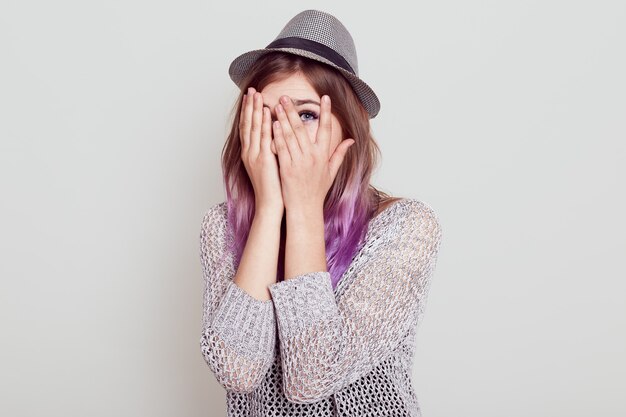 Mujer joven caucásica se asoma a través de los dedos, se cubre la cara con las manos, vestida con sombrero, teme algo, mira tirar los dedos, aislado sobre fondo gris.