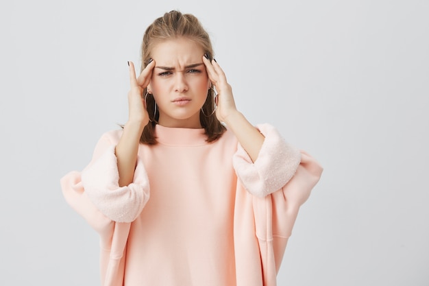 La mujer joven caucásica linda se vistió en la sudadera rosada que fruncía el ceño su cara que parecía cansada que sostenía las manos en las sienes debido al cansancio o al dolor de cabeza. Mujer joven con expresión triste y dolor de cabeza.