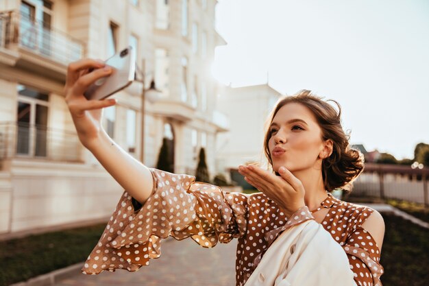 Mujer joven caucásica refinada con teléfono para selfie en día de otoño. Tiro al aire libre de glamorosa modelo femenina en ropa marrón enviando beso al aire.