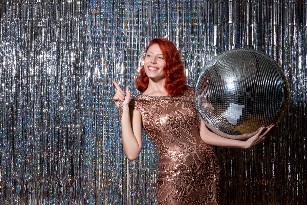 Mujer joven celebrando el año nuevo en la fiesta sosteniendo una bola de discoteca en cortinas cortinas brillantes