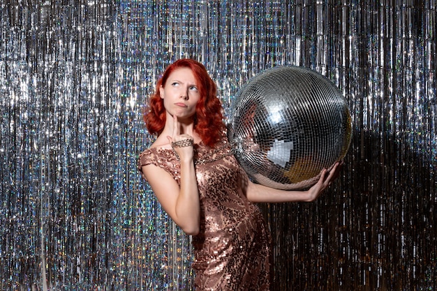 Mujer joven celebrando el año nuevo en la fiesta sosteniendo una bola de discoteca en cortinas cortinas brillantes