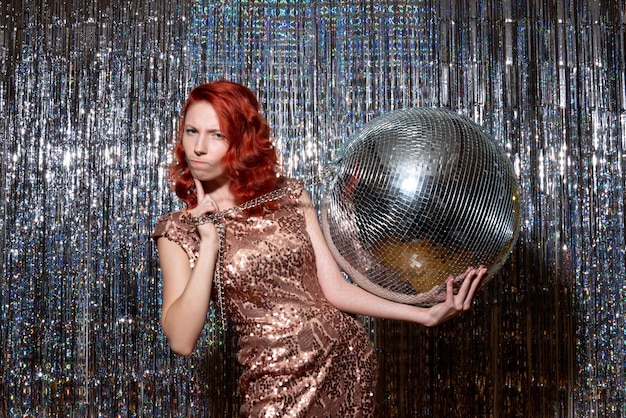 Mujer joven celebrando el año nuevo en la fiesta sosteniendo una bola de discoteca en cortinas cortinas brillantes