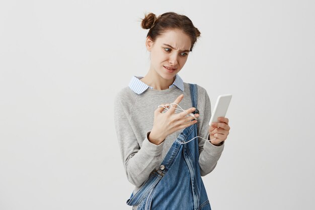 La mujer joven con el ceño fruncido que usa el teléfono móvil no puede escuchar música debido a los auriculares descifrados. Chica de moda que tiene pocos problemas con sus auriculares. Situación y solución.