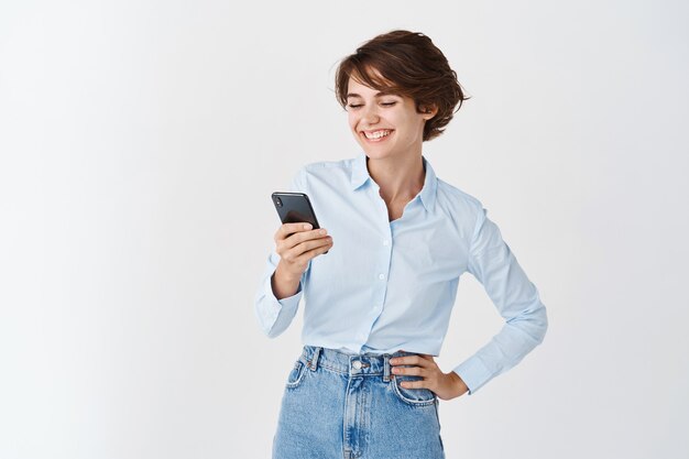 Mujer joven ceo profesional leyendo la pantalla del teléfono y sonriendo, usando el teléfono inteligente en la pared blanca