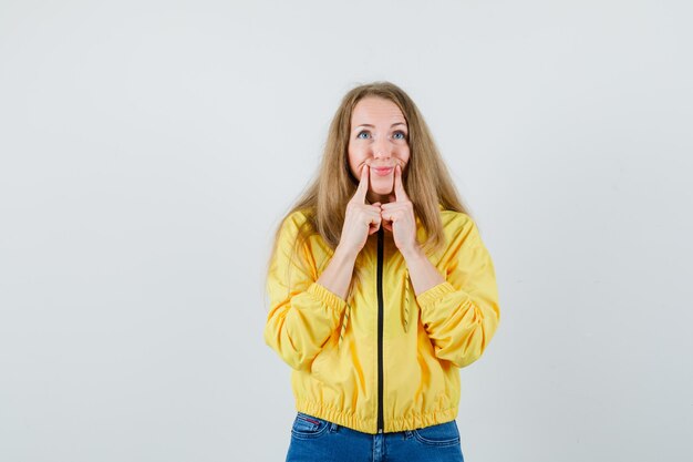 Mujer joven en chaqueta de bombardero amarilla y jean azul apuntando a la boca con los dedos índices y hinchando las mejillas y mirando serio, vista frontal.