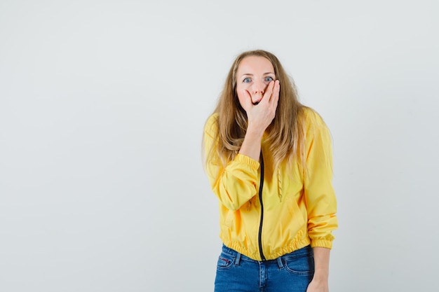 Mujer joven en chaqueta de bombardero amarilla y jean azul cubriendo su boca y mirando sorprendido, vista frontal.