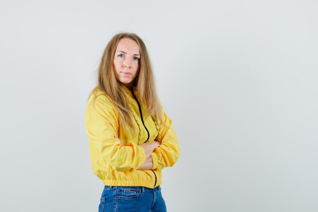 Mujer joven en chaqueta de bombardero amarilla y jean azul de pie con los brazos cruzados y posando a la cámara y mirando encantador, vista frontal.
