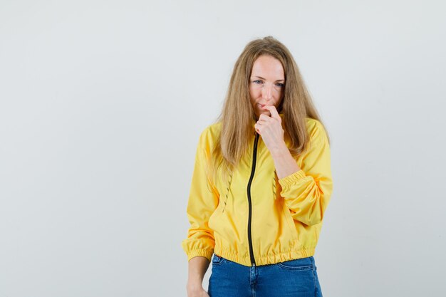 Mujer joven en chaqueta de bombardero amarilla y jean azul de pie en pose de pensamiento y mirando pensativo, vista frontal.