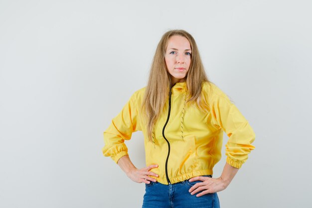 Mujer joven en chaqueta de bombardero amarilla y jean azul tomados de la mano en la cintura y posando en la cámara