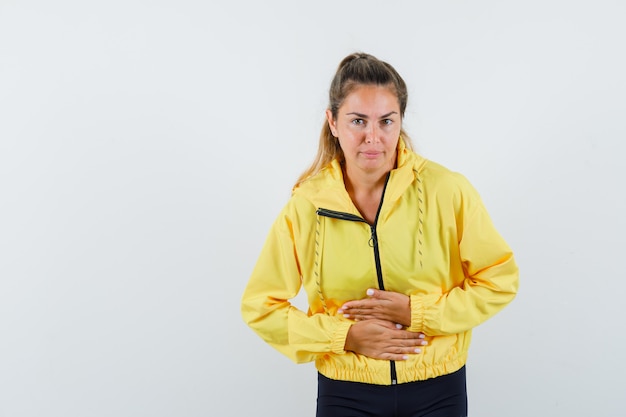 Mujer joven en chaqueta de bombardero amarilla y pantalón negro poniendo las manos en el vientre y con dolor de estómago y mirando serio