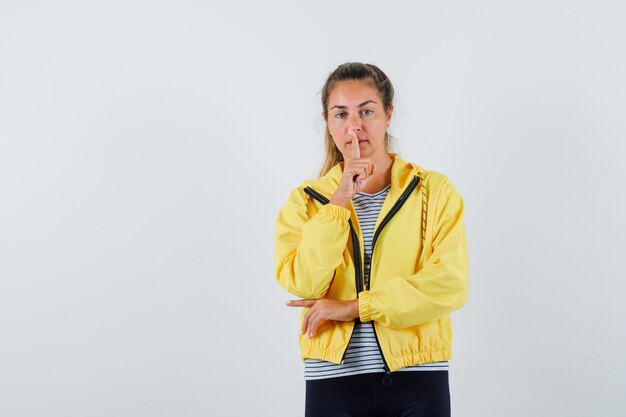 Mujer joven en chaqueta, camiseta mostrando gesto de silencio y mirando confiado, vista frontal.