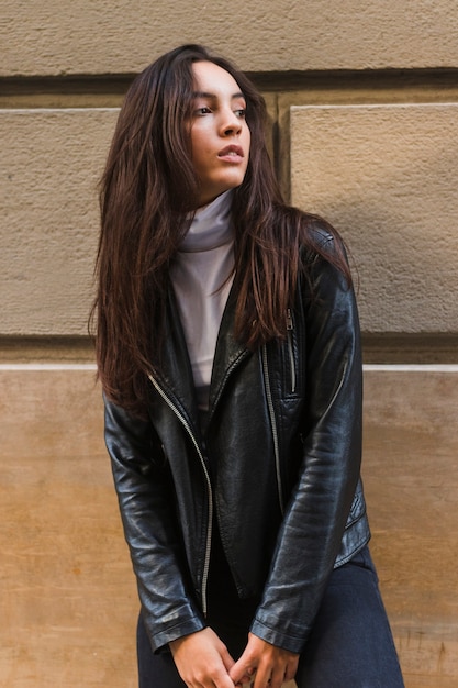 Mujer joven en la chaqueta de cuero negra que se opone a la pared que mira lejos