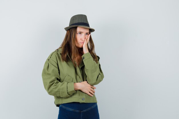 Mujer joven en chaqueta, pantalón, sombrero sosteniendo la mano en la mejilla y mirando molesto, vista frontal.