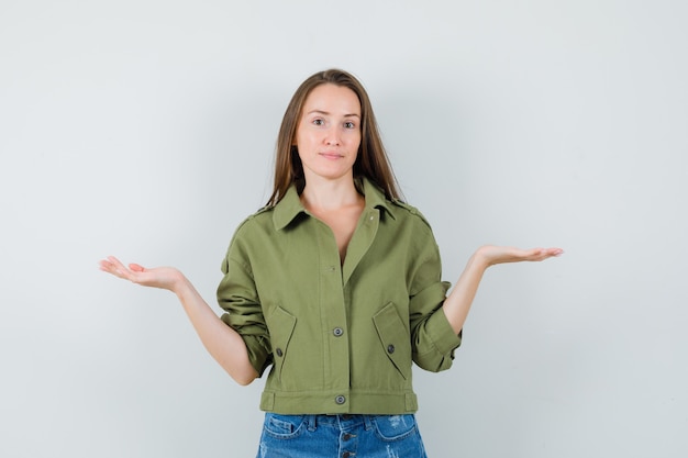 Mujer joven en chaqueta, pantalones cortos presentando o comparando algo y mirando confiado, vista frontal.