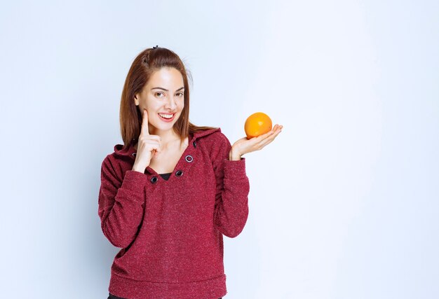 Mujer joven en chaqueta roja sosteniendo una naranja y parece pensativa