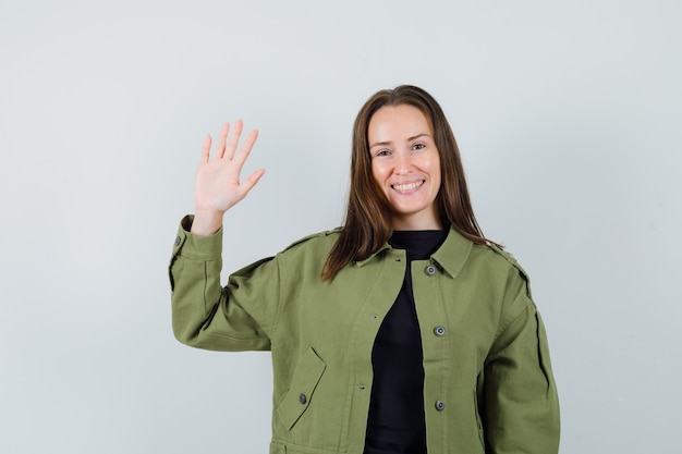 Mujer joven en chaqueta verde agitando la mano para saludar y mirar alegre, vista frontal.