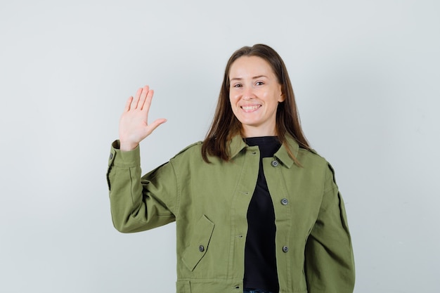 Mujer joven en chaqueta verde agitando la mano para saludar y mirar contento, vista frontal.
