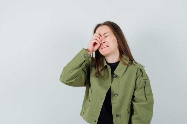 Mujer joven en chaqueta verde frotándose los ojos mientras llora y mira triste, vista frontal.