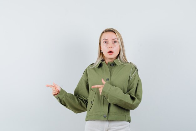 Mujer joven en chaqueta verde, jeans apuntando a un lado y mirando emocionado, vista frontal.