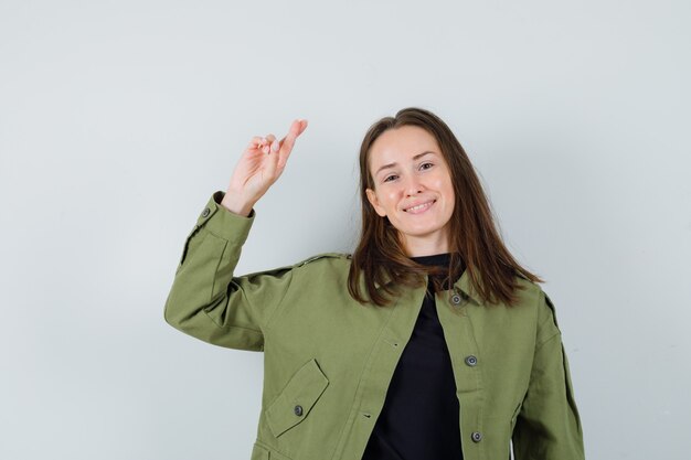 Mujer joven con chaqueta verde levantando la mano con los dedos cruzados y mirando satisfecho, vista frontal.