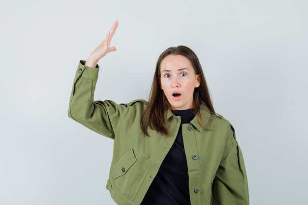 Mujer joven en chaqueta verde levantando la mano con la palma abierta y mirando agresivo, vista frontal.