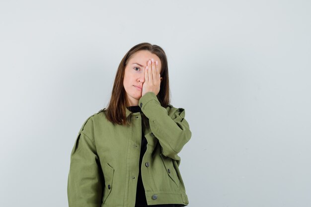 Mujer joven en chaqueta verde con la mano en la cara y mirando oculta, vista frontal.