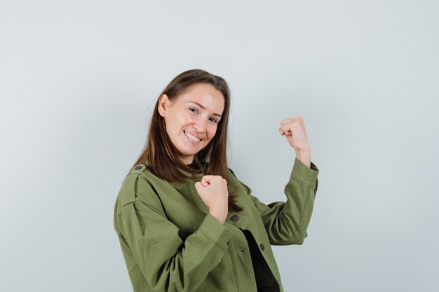 Mujer joven en chaqueta verde mostrando gesto de ganador y mirando alegre, vista frontal.